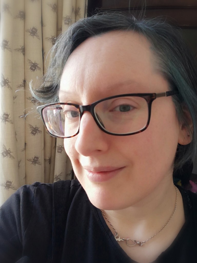 Image shows Sophie, a light skinned woman with dark grey hair. She is wearing dark framed glasses, a black top, and a silver chain. She is looking at the camera and smiling. In the background are cream coloured curtains. 